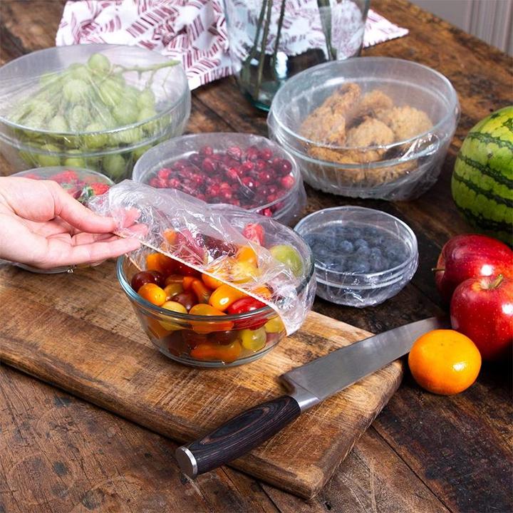 Verschiedene Schüsseln mit Obst und Keksen, mit PlasticWrap Frischhaltehauben abgedeckt, auf Holztisch